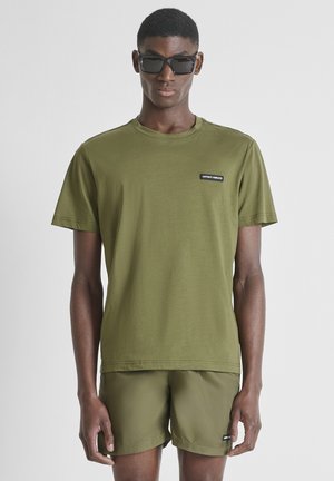 Man draagt olijfgroen T-shirt en korte broek met kleine zwarte labels, zwarte zonnebril, staand tegen een effen lichte achtergrond.
