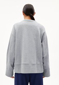 Eine Person mit dunklem Haar, das zu einem Zopf gebunden ist, trägt einen übergroßen hellgrauen Pullover und marineblaue Hosen, gesehen von hinten.