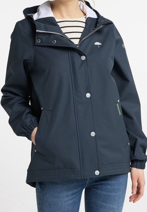 Femme portant une veste imperméable bleu marine avec boutons-pression, fermeture éclair et poignets élastiques, par-dessus une chemise rayée et un jean bleu.
