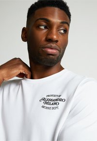 Valkoinen puuvillapaita, jossa on mustilla brodeeratuilla teksteillä "PROPERTY OF ALESSANDRO MILANO ARCHIVE DEPT." rintamalla; pehmeä rakenne, pyöreä kaula-aukko.