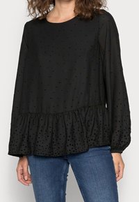 Femme portant un blouse noire à manches longues transparentes, avec un motif à pois et un ourlet à volants, assortie à un jean en denim bleu.