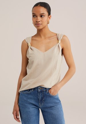 Femme aux cheveux foncés attachés, portant un haut beige sans manches avec des détails de bretelles dorées et un jean bleu, une main dans la poche, fond neutre.