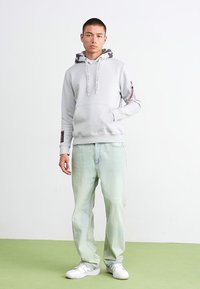 Lys grå hettegenser med en hette i kamuflasjemønster, sidelommer med glidelås, sammen med lysegrønne jeans og hvite sneakers, står på en grønn overflate.