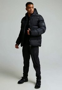 Melna pūkaina jaka ar kapuci, uz pleca ir logotipa uzlīme un priekšpusē rāvējslēdzējs. Valkāta ar melnām biksēm un sporta apaviem.