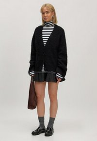 Schwarze, flauschige Strickjacke mit V-Ausschnitt und Knopfverschluss, über einem gestreiften Rollkragenpullover getragen. Kombiniert mit einem schwingenden Minirock und schwarzen Slippern.