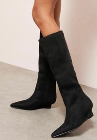 Fekete velúr térdige érő csizmák hegyes orral és alacsony wedge sarokkal. Oldalán cipzárral és sima felülettel rendelkezik.