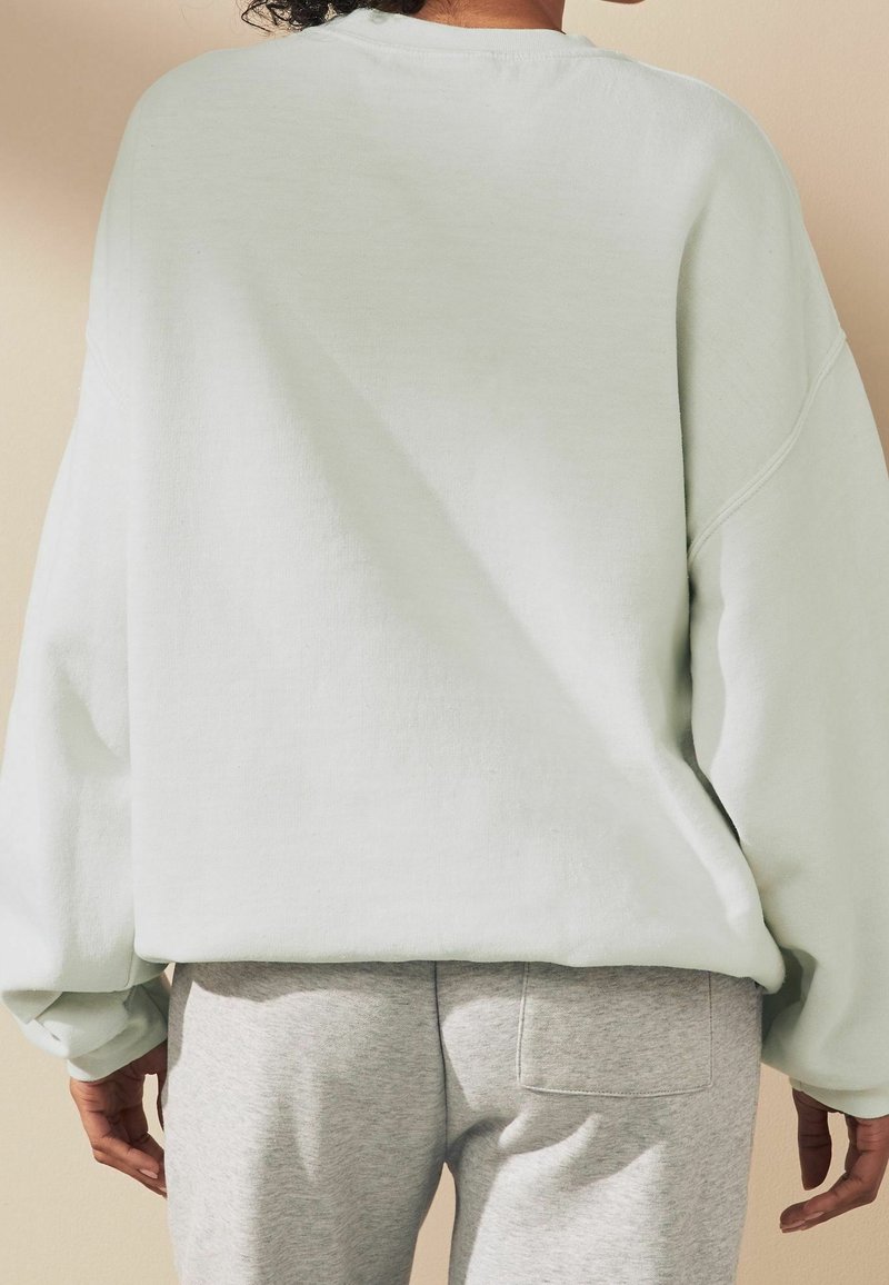 Felpa oversized verde chiaro con spalline scese, polsini a coste e una finitura morbida e texturizzata, abbinata a pantaloni felpati grigi con tasca posteriore.