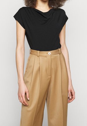 Vrouw draagt een zwarte top met korte mouwen, ingestopt in high-waisted, wijde camel-kleurige broek met een getailleerde, met knopen sluitende tailleband.