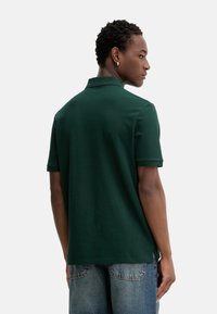 Polo de color verde oscuro con mangas cortas, cuello y una textura suave. Llevado con jeans de mezclilla azul claro, mostrando la vista por detrás.