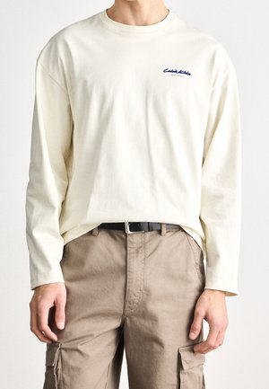 Personne portant un t-shirt crème à manches longues avec un petit logo bleu sur la poitrine, un pantalon cargo beige et une ceinture noire, debout devant un fond uni.