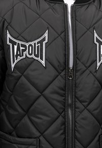 Čierna prešívaná bomber bunda s nášivkami TAPOUT. Obsahuje zips vpredu a dve bočné vrecká, s hladkou textúrou a odolným materiálom.