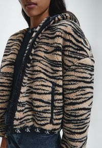 Kurtka z futra syntetycznego w wzór w paski tygrysa w odcieniach beżu i czerni. Posiada zamek błyskawiczny, kieszenie boczne oraz taśmę z logo na dolnym brzegu.