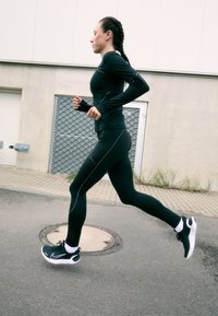 Zwarte sportieve long-sleeve shirt en leggings met reflecterende accenten, gecombineerd met zwart-witte hardloopschoenen, gedragen door een persoon die buiten aan het hardlopen is.