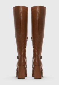 Botas altas de cuero marrón con tacones en bloque y cremalleras laterales, vistas desde atrás sobre un fondo blanco liso.