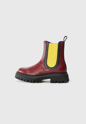 Red leather ankle boot with a chunky black sole, featuring yellow elastic side panels and a pull tab at the back. Textured finish.