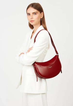 Jeune femme en blazer blanc portant un sac à bandoulière rouge Lancaster en cuir, les bras croisés, sur fond blanc.