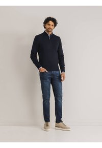 Marineblauer Zip-Pullover mit langen Ärmeln, kombiniert mit blauen Jeans und beigen Sneakers, bietet eine schmale Passform und eine strukturierte Oberfläche.