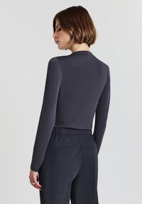 Donna con i capelli corti castani che indossa una maglietta attillata grigia scura a maniche lunghe e pantaloni scuri a vita alta, mostrata di spalle su uno sfondo bianco.