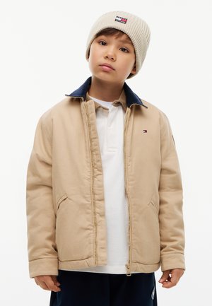 Enfant portant une veste beige avec col bleu marine, chemise blanche, pantalon bleu marine et bonnet en tricot beige avec logo, debout sur fond blanc.