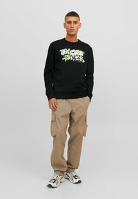 Sudadera negra con texto gráfico en verde y blanco, pantalones cargo beige con bolsillos laterales y zapatillas grises con acentos en negro y blanco.