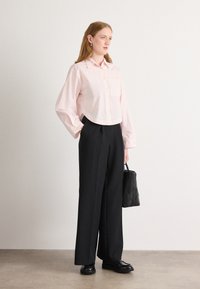Camicetta corta rosa chiaro con colletto a bottoni e taschino sul petto, abbinata a pantaloni ampi neri. Porta una piccola borsa a tracolla nera trapuntata.