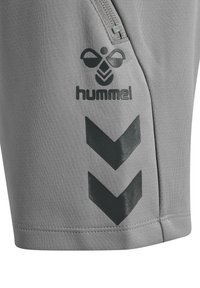 Close-up van grijze sportkledingstof met zwart Hummel-logo, ritssluitingdetail en twee chevronstrepen.