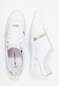 Bílé tenisky se zlatými akcenty, s perforovaným vzorem, zavazováním na tkaničky a texturovanou podrážkou. Logo viditelné na stélce.