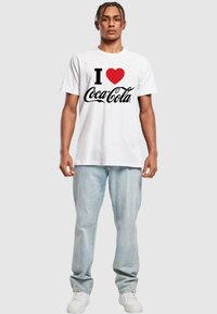 Weißes Baumwoll-T-Shirt mit einem schwarzen "Coca-Cola"-Logo und einem roten Herz. Kombiniert mit hellblauen Jeans und weißen Sneakers.