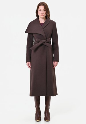 Donna che indossa un lungo cappotto marrone scuro con cintura e ampio colletto, insieme a stivali marroni al ginocchio, in piedi davanti a uno sfondo chiaro e semplice.
