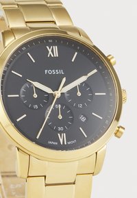 Montre dorée avec un cadran noir, trois sous-cadrans et affichage de la date. Dispose d'un bracelet en acier inoxydable et d'un mouvement à quartz.