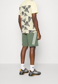 Keltainen ja musta graafinen t-paita, jossa on teksturoitu selkä, yhdistettynä vihreisiin shortseihin, joilla on valkoiset raidat, ja harmaisiin lenkkarit joissa on oransseja yksityiskohtia.