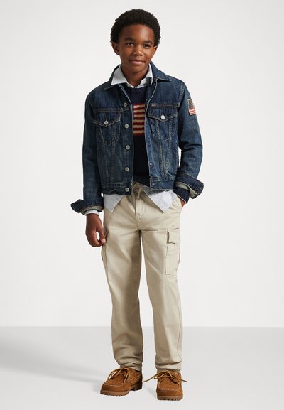 Τζάκετ από τζιν με μπαλώμα με την αμερικανική σημαία, φορεμένο πάνω από ένα ριγέ πουλόβερ και πουκάμισο με γιακά, σε συνδυασμό με μπεζ cargo παντελόνι και καφέ παπούτσια.