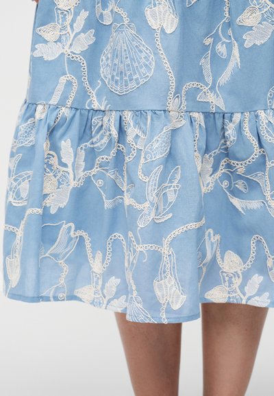 Jupe bleue avec des motifs brodés blancs sur le thème de la mer, comprenant des coquillages, des poissons, des algues et des chaînes, portée sur des jambes visibles en dessous de l'ourlet.