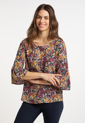 Vrouw met lang bruin haar die glimlacht, draagt een bloes met bloemenprint en klokmouwen en een donkere broek, staand tegen een effen witte achtergrond.