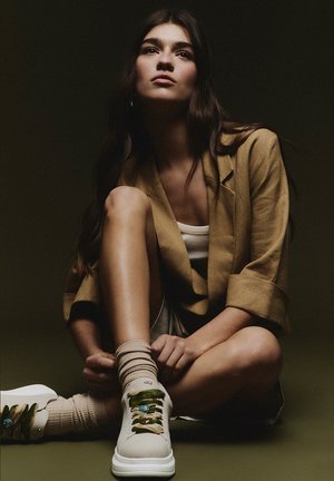 Mujer joven con cabello largo sentada en el suelo, vestida con blazer beige, camiseta blanca, pantalones cortos, calcetines beige y zapatillas blancas con cordones decorativos.