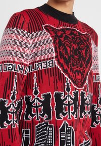 Gestrickter Pullover mit kräftigen roten und schwarzen Mustern, darunter eine Bärengrafik und architektonische Motive, mit einem gerippten schwarzen Rundhalsausschnitt.