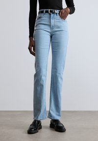 Svijetloplave traperice ravnog kroja s visokim strukom, s petljama za remen i klasičnim dizajnom s pet džepova. Nosit s crnim remenom i cipelama.