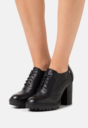 Chaussures oxford en cuir noir avec un talon épais, présentant des détails de brogue, une surface texturée et un design à lacets. Semelle en caoutchouc avec une bonne adhérence.