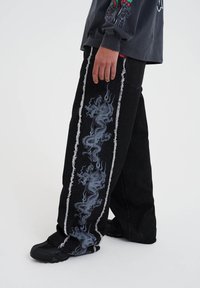 A person wearing black jeans with dragon graphics on the side stands on a plain background, paired with a dark long-sleeve shirt.