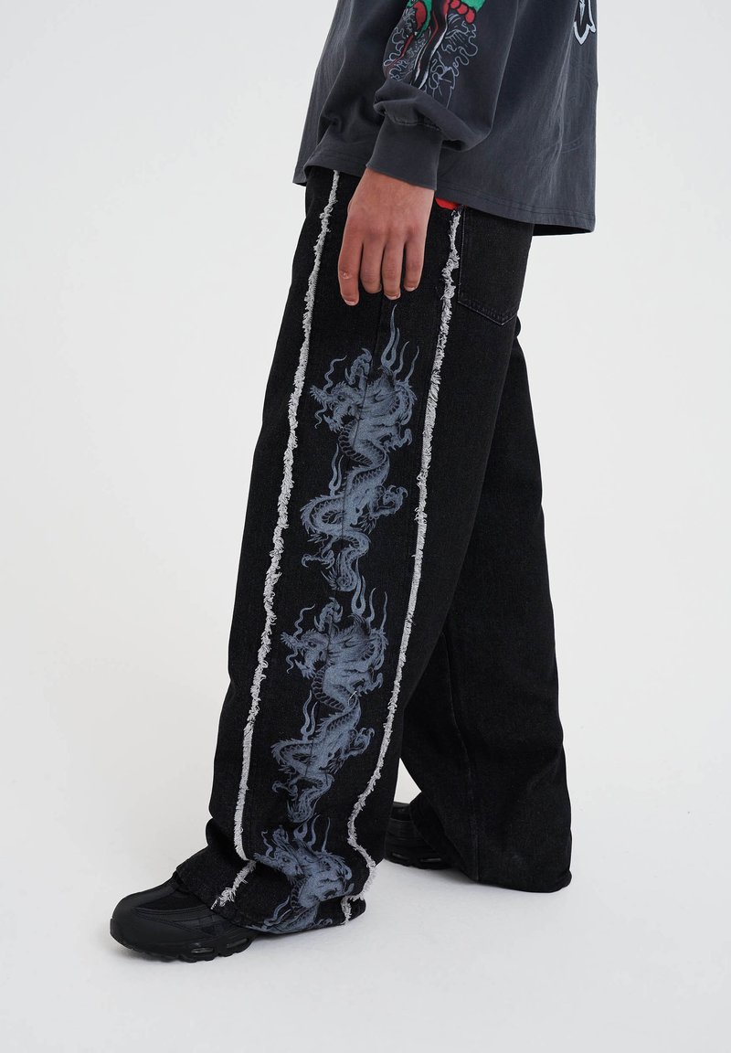 A person wearing black jeans with dragon graphics on the side stands on a plain background, paired with a dark long-sleeve shirt.