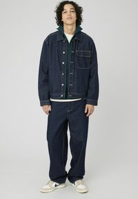 Veste en denim foncé avec fermeture à boutons et poche, associée à un hoodie zippé vert foncé et un jean ample foncé. Baskets blanches avec des accents.