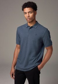 Camisa polo azul oscura de manga corta, con cuello y un logo sutil en el lado izquierdo del pecho, combinada con pantalones negros. Textura de tela suave.