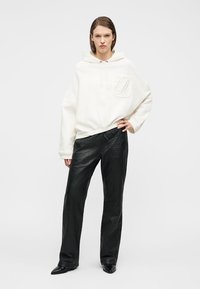 Γυναίκα που ποζάρει φορώντας μια λευκή φούτερ με μεγάλο μέγεθος με το γράμμα "EA" στην τσέπη, μαύροι υφασμάτινοι παντελόνες με ευρύ πόδι και μαύρα παπούτσια με μυτερή μύτη, μπροστά σε ένα απλό φόντο.