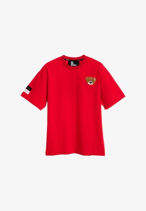 Mujer rubia con camiseta roja extragrande con un gran dibujo de un osito en la espalda, pantalones cortos beige, calentadores de piernas y zapatos, de pie mirando hacia atrás.