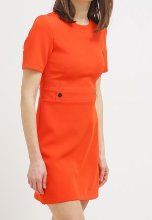 Woman wearing a short-sleeve, knee-length bright orange dress with black button details at the waist, standing against a white background.