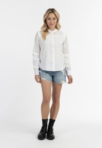Witte blouse met een golvende kraag en eyelet-details, gecombineerd met versleten denim shorts en zwarte veterschoenen.