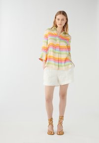 Färgglad blus med zigzag-mönster, krage och trekvartslånga ärmar, kombinerad med vita linnebyxor och gula sandaler med remmar.
