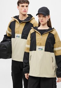 Δύο άτομα φορούν πουλόβερ με τμήματα σε μαύρο, μπεζ και καφέ χρώμα. Τα πουλόβερ διαθέτουν μισό φερμουάρ, κουκούλα και ταμπέλα με λογότυπο.