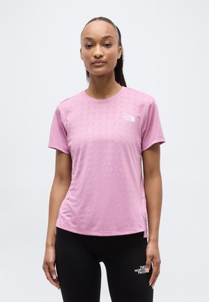 Moteris, vilkinti šviesiai violetinę trumpomis rankovėmis palaidinę ir juodas sportines kelnes, ant kurių abiejų matyti The North Face logotipas, stovinti prieš baltą foną.