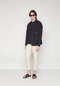 Melna zip-up jakna ar divām krūšu kabatām virs melnas krekla, apvienota ar gaiši bēšām sašaurinātām biksēm un melnām slaidām sandālēm.